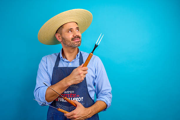 istock Chef wearing straw hat and apron holding barbecue fork looking up on blue background 2235432144