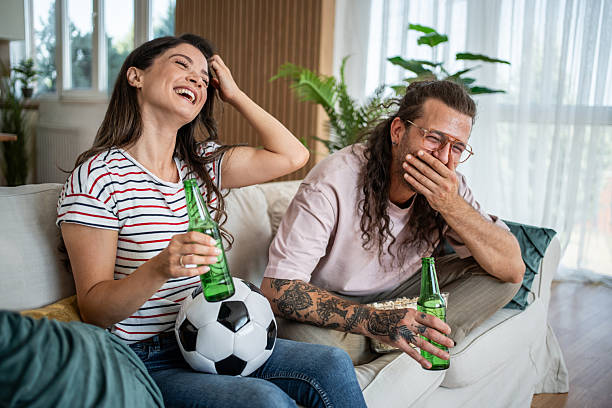 istock Couple laughing while watching football and drinking beer at home 2235432267