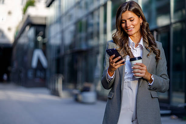 istock Young businesswoman is using smartphone and holding coffee while walking outside office building 2235452241