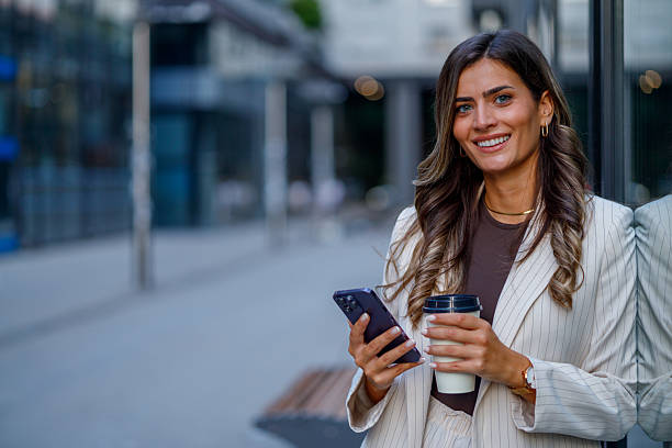istock Young businesswoman enjoying coffee while using her smartphone outdoors, smiling confidently in a modern city setting 2235452584