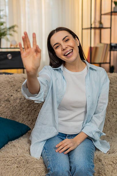 istock Young woman sitting on sofa smiling cheerfully waving hand hello greeting friendly welcoming gesture 2235454709