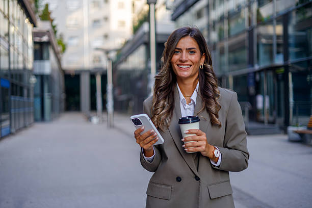 istock Smiling businesswoman using smartphone and holding disposable coffee cup while walking outside office building 2235457173