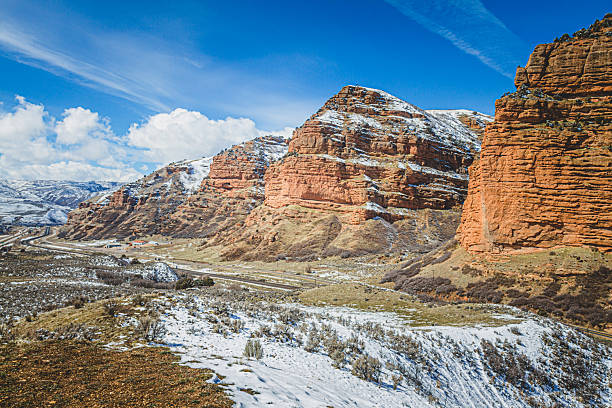 Majestic red rock formations rise above a snow-covered landscape, with patches of greenery visible. The bright blue sky adds to the scenic beauty of this rural location.