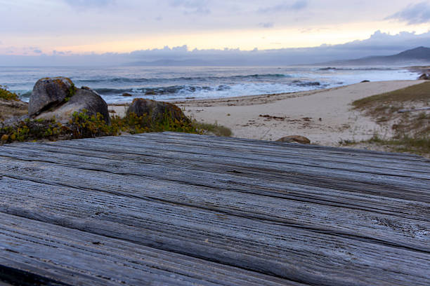 A wooden table sits on a beach near the ocean. The table is empty and the beach is rocky. The sky is cloudy and the sun is setting