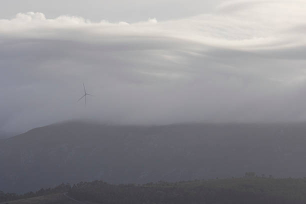 A wind turbine is visible in the sky above a mountain range. The sky is cloudy and the wind is blowing