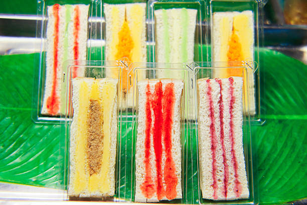 Horizontal image of Thai street food sandwiches filled with strawberry, pandan cream, pork floss, and blueberry. The soft white bread highlights colorful layers, arranged on banana leaf inside a transparent plastic container. Each sandwich slice showcases a blend of sweet and savory flavors, representing Thailand’s vibrant food culture. Perfect for concepts of fusion food, Asian cuisine, and modern Thai snacks.