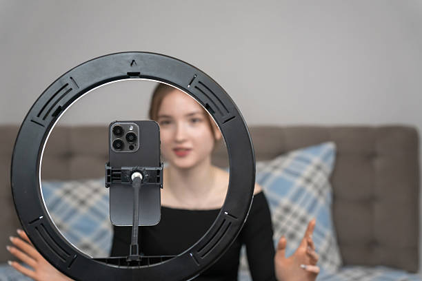 istock Young girl vlogger recording video with smartphone using ring light while sitting on bed at home, creating engaging content for social media platforms. Focus on the ring lamp 2235497674