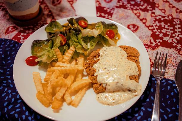 A first-person, eye-level view of a person eating a plate of Nasi Campur, a traditional Indonesian mixed rice dish, at a rustic restaurant. The plate sits on a table covered with a vibrant red and white Indonesian batik tablecloth.
