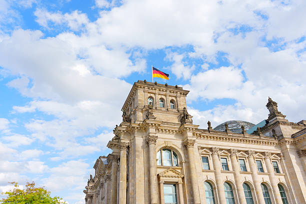 Berlin, Germany - August 20, 2025: Majestic view of the Reichstag building featuring German flag against a blue sky with fluffy clouds.