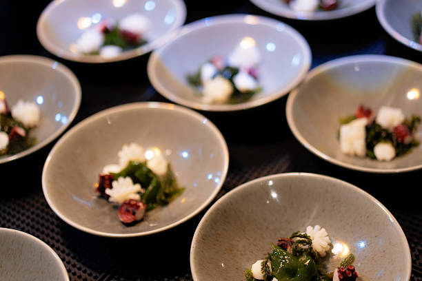 In a restaurant kitchen, plating a series of gourmet appetizers. Each identical gray bowl on the counter is being filled with a small portion of seafood and sea greens.