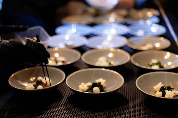 A chef, wearing black gloves, is captured in a wide shot in a restaurant kitchen, actively plating a series of gourmet appetizers. Each identical gray bowl on the counter is being filled with a small portion of seafood and sea greens.