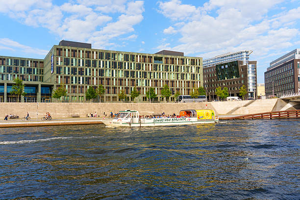 Berlin, Germany - August 20, 2025: Boat cruising on the Spree River with modern buildings lining the riverbank on a sunny day.