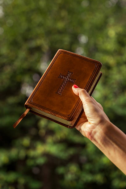 Close-up of a hand lifting a brown leather-bound Bible with embossed cross, set against a blurred green outdoor background.