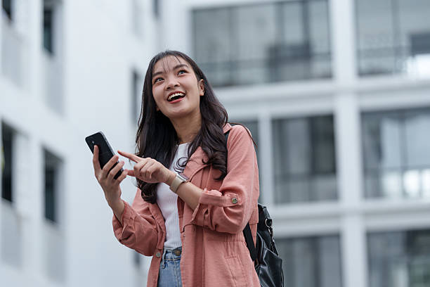 istock Young asian woman with a backpack smiling and looking up while using her smartphone in front of a modern building, enjoying the vibrant atmosphere of urban life 2235504779