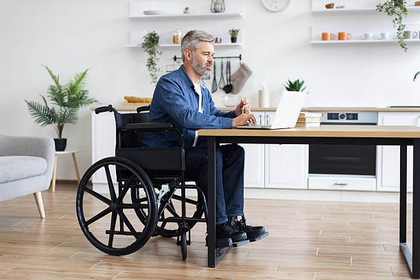 istock Man in wheelchair working on laptop in bright modern home kitchen 2235505175