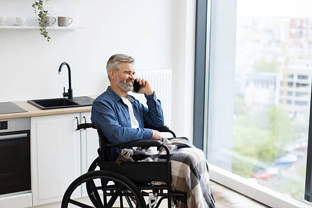 istock Man in wheelchair by kitchen using phone next to large window 2235505213