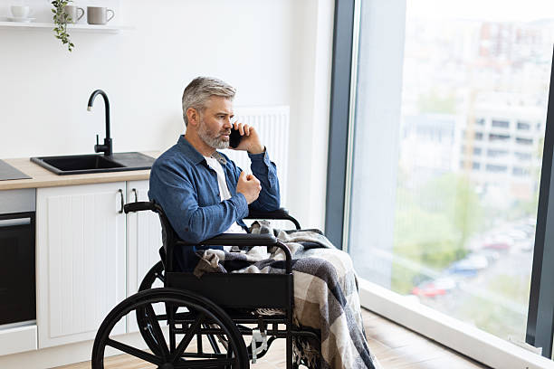 istock Man in wheelchair by kitchen using phone next to large window 2235505215