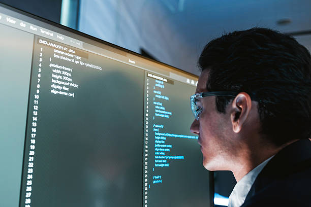 istock Software developer wearing glasses focuses on code displayed on a large monitor in a modern office, reviewing and debugging source files on a wide screen workstation. 2235509511