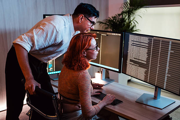 istock Two colleagues work together at a dual-monitor desk in a modern office, reviewing code or data at night. One stands and guides while the other types, screens glowing in low light. 2235509516