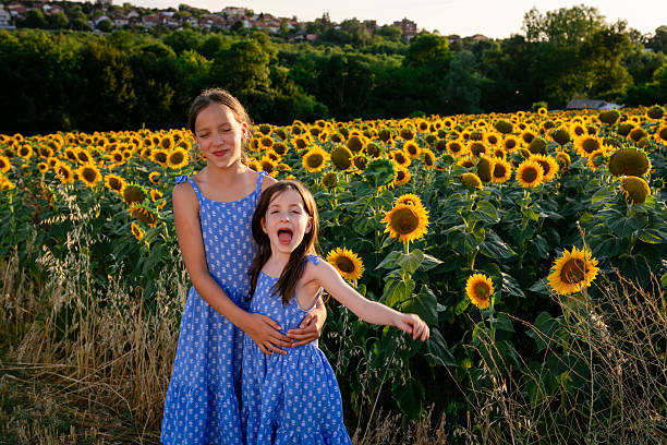 Two sisters wearing blue dresses are enjoying a moment of joy and laughter in a beautiful field of sunflowers at sunset