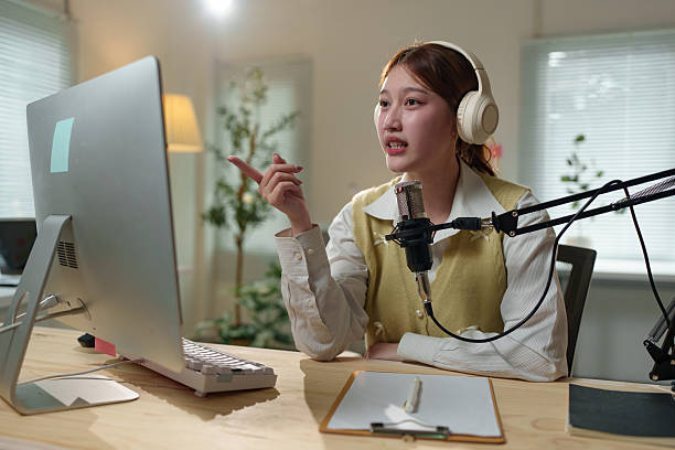 istock Young asian female podcaster wearing headphones and speaking into a microphone while pointing at the computer screen during a home recording session 2235510795