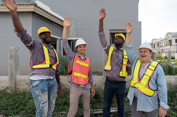 istock Four construction worker raising their hand high marking a victorious moment, stand outside a building site. They wear hardhat and safety vest. 2235513142