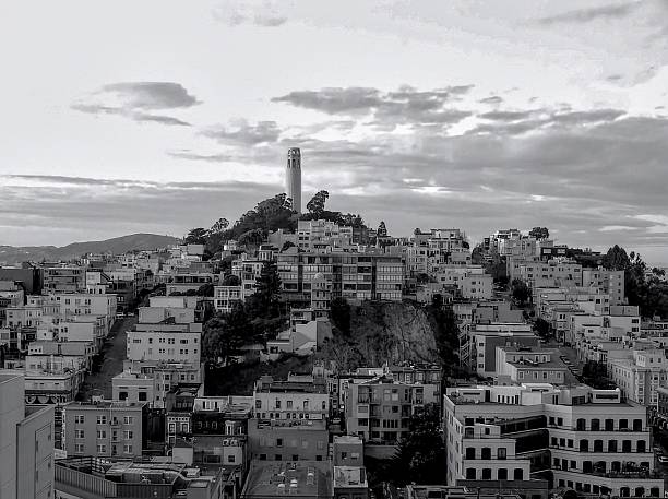 Black and white view of San Francisco's Coit Tower atop Telegraph Hill with surrounding cityscape under a cloudy sky.