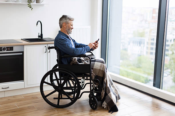 istock Man in wheelchair using smartphone in modern apartment with large windows 2235520230
