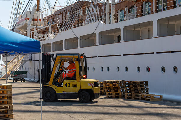 Venice, Italy, August 11, 2025: A dockworker operates a yellow forklift to move wooden pallets beside a large white cruise ship at a port on a sunny day.