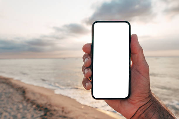 istock Close-up of hand holding smartphone with blank white screen on beach at sunset, app or web design mockup. 2235521708