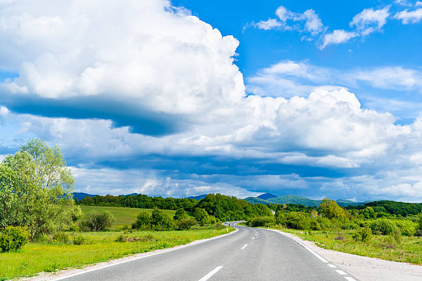 istock Empty Curved Road Through Green Fields with Trees Under Bright Blue Sky and White Clouds in Countryside Scenic Landscape View 2235524571