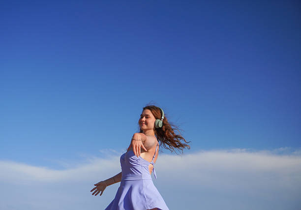 Young woman in light purple dress dances outdoors under blue sky with headphones. Clouds and open space in background.