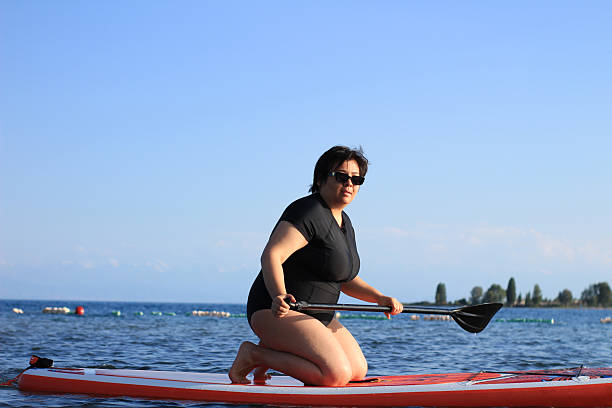 Woman in black swimsuit kneels on paddleboard, holding paddle, on calm lake, clear blue sky.
