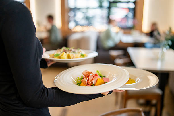 istock Restaurant server in black uniform carrying three plated meals, including salad, pasta, and polenta, in a modern dining setting with blurred guests and wooden furniture 2235534157