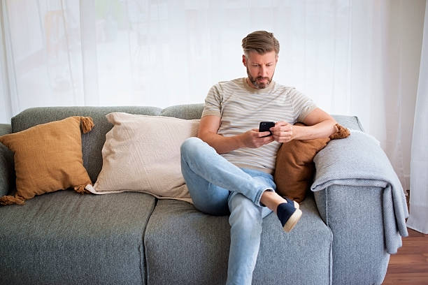 istock Middle aged man sitting on the couch at home and using his cell phone 2235535286