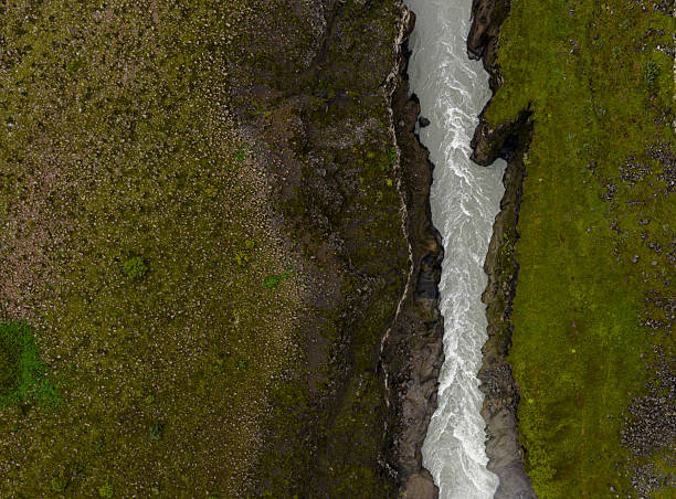 A narrow river flows swiftly through a rugged canyon in Iceland, with steep dark walls and vibrant green moss contrasting the rocky terrain.