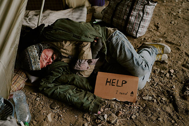 Middle aged Caucasian man sleeping on ground outdoors near tent holding cardboard sign reading help I need, wearing layered clothing, surrounded by personal belongings and debris