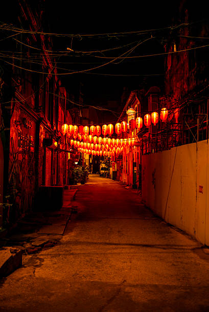 A nighttime scene of a narrow alleyway illuminated by a string of glowing red lanterns overhead. The warm light reflects off the old buildings and ground, creating a festive yet mysterious atmosphere reminiscent of traditional celebrations or Chinatown streets.