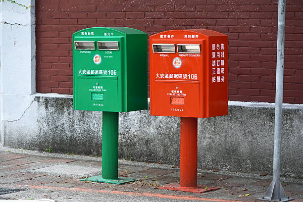 Taiwan - Sep 14, 2025: Two Taiwan Post mailboxes, green on left and red on right, with cylindrical posts display collection times and postal codes against red brick wall background.