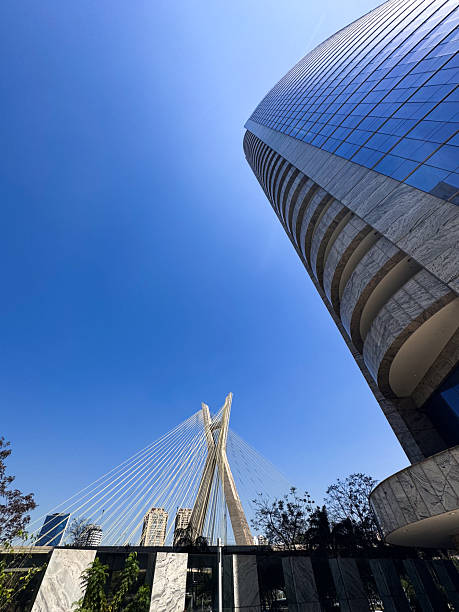 A modern architectural scene featuring a towering skyscraper and the iconic Ponte Estaiada bridge against a bright blue, cloudless sky. The composition highlights São Paulo's urban design and engineering excellence, blending the structural artistry of the bridge with the reflective glass facade of the building.