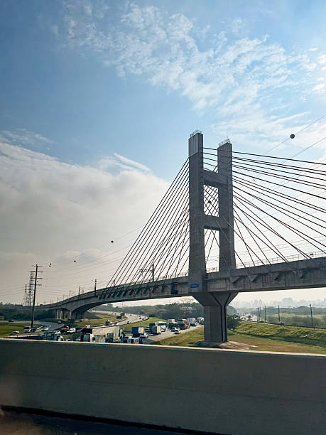 An iconic cable-stayed sky Guarulhos train  bridge spans across a highway under a tranquil, cloudy sky. The scene captures the blend of modern infrastructure and everyday activity, with cars and trucks traveling beneath. This urban landmark reflects the essence of innovation and the importance of connectivity in city life.