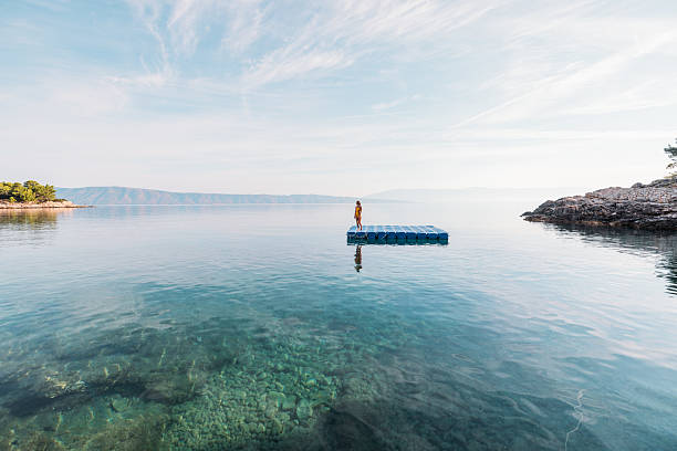 Young woman in yellow swimsuit standing on a floating dock in calm sea water near rocky shore. Solo adult enjoying peaceful moment on water platform. Person relaxing by the ocean.