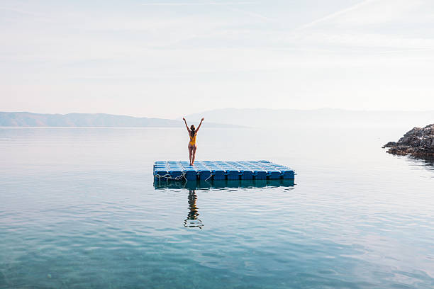 Young woman in yellow swimsuit stands with arms raised on floating dock in clear calm sea near rocky shore. Person enjoying solitude and peaceful water landscape. Relaxation and freedom.
