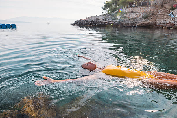 Young woman in yellow swimsuit floating on back in clear calm sea near rocky shore. Relaxed adult enjoying peaceful water and sunlight. Leisure and tranquility in nature.