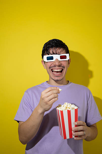 istock Man with 3D glasses eating popcorn from a cardboard box on yellow background with copy space 2235580917
