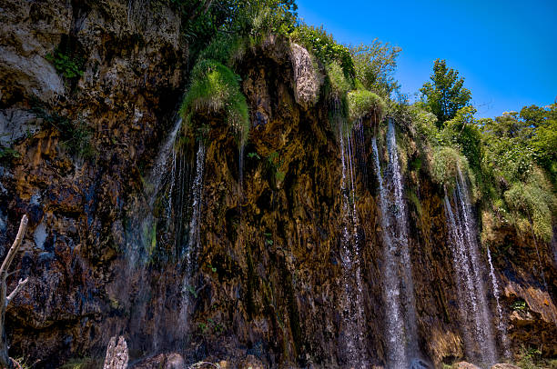 Plitvice Lakes, Croatia – July 21, 2025: Water spills over moss-covered cliffs in a series of thin cascading waterfalls beneath a bright summer sky.
