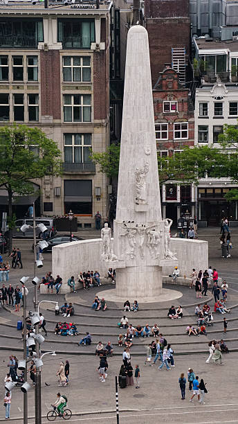 editorial photo, taken on August 17, 2025, in Amsterdam, captures the National Monument at Dam Square. The white stone obelisk, dedicated to the victims of World War II, stands as the focal point of the composition. Surrounding the monument are numerous people, some sitting, walking, or taking photos. In the background, historic and modern buildings provide architectural context