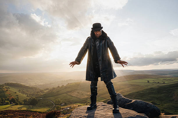 Individual wearing a coat and top hat poses on a rock, surrounded by a beautiful landscape of rolling hills and a colorful sunset sky, creating a striking silhouette.