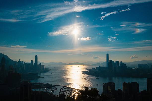 A stunning view of Hong Kong's skyline with a bright sun over Victoria Harbour, reflecting on the water.