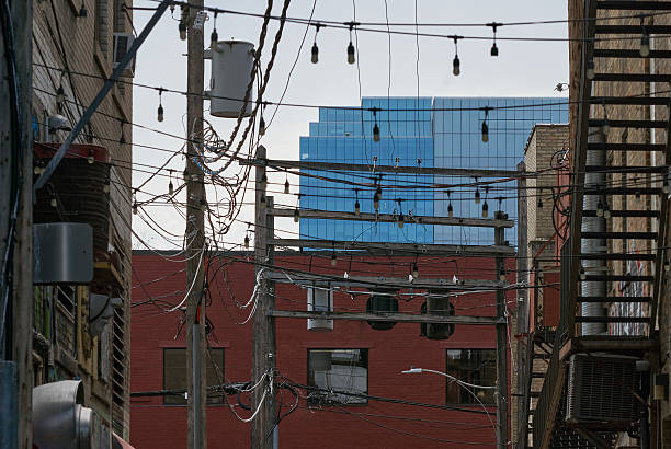 A city alley in downtown Saskatoon, Canada, filled with tangled overhead utility wires, wooden poles, and hanging light bulbs. In the background, a modern glass office tower rises, creating a strong contrast between old infrastructure and contemporary architecture. The image highlights the complexity of urban electrical systems and layered cityscapes.